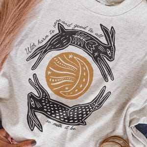May include: A light gray crewneck sweatshirt featuring a black and mustard yellow graphic of two rabbits circling a crescent moon. The text reads, With harm to none and good to all, so mote it be, printed in a black script font across the design.