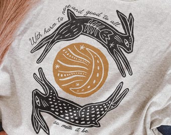 May include: A light gray crewneck sweatshirt featuring a black and mustard yellow graphic of two rabbits circling a crescent moon. The text reads, With harm to none and good to all, so mote it be, printed in a black script font across the design.