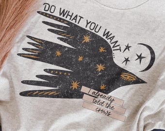 May include: A light gray crewneck t-shirt featuring a black graphic of a flying crow with gold stars and a crescent moon. The text reads DO WHAT YOU WANT... and I already told the crows. The design has a distressed, weathered texture.