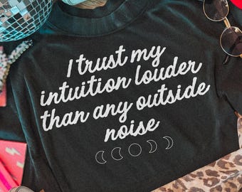 May include: A black long-sleeve shirt featuring the white text I trust my intuition louder than any outside noise and a series of moon phase icons. The shirt is surrounded by a disco ball, light-colored sneakers, sunglasses, and a leopard print fabric.