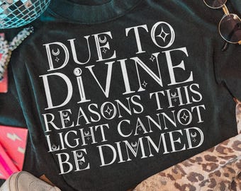 May include: A black sweatshirt featuring white text that reads DUE TO DIVINE REASONS THIS LIGHT CANNOT BE DIMMED, accompanied by a silver disco ball, light-colored high-top sneakers, round sunglasses, a leopard print fabric, and a pink card.