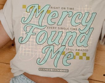 May include: A cream-colored short-sleeve t-shirt featuring the text "Mercy Found Me" in large, light blue script. Additional text includes "RIGHT ON TIME," "EVERY SINGLE TIME," "STILL AMAZED," and "AMAZING GRACE CLUB" over a grid and checkerboard pattern.