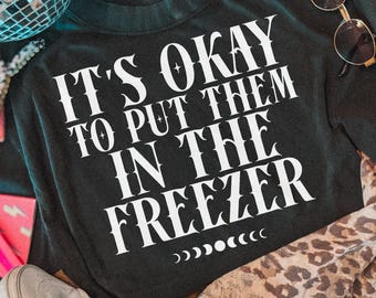May include: A black sweatshirt featuring white text that reads IT'S OKAY TO PUT THEM IN THE FREEZER with a moon phase graphic below. The garment is surrounded by a disco ball, light-colored sneakers, round sunglasses, and a leopard print fabric.