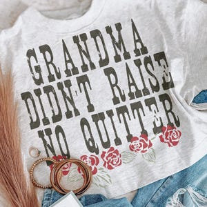 May include: A light gray heathered t-shirt featuring the dark green distressed text "GRANDMA DIDN'T RAISE NO QUITTER" printed across the chest, accented by a row of red roses with green leaves positioned below the lettering.