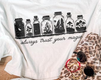 May include: A white t-shirt featuring a black graphic of seven glass jars containing various items like crystals, plants, and mushrooms, with the text "always trust your magic" printed underneath in a cursive font.
