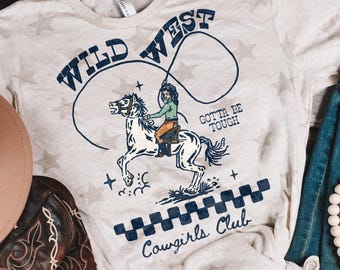 May include: A light grey t-shirt with an all-over star pattern featuring a graphic of a person riding a horse with a lasso. The text reads WILD WEST, GOTTA BE TOUGH, and Cowgirls Club, accompanied by a dark blue checkered border design.