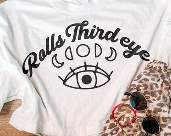 May include: A white short-sleeve t-shirt with black text that reads Rolls Third eye above a graphic of moon phases and an eye. The shirt is paired with leopard print shorts, a pair of black sunglasses, and two beaded bracelets.