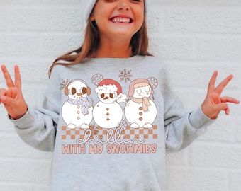 Puede incluir: Una sudadera gris claro de cuello redondo con un gráfico de tres muñecos de nieve con accesorios de invierno, como orejeras, un gorro de Papá Noel y un gorro de trampero. Debajo hay un patrón de cuadros y el texto "CHILLIN WITH MY SNOWMIES."