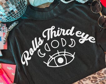 May include: A black long-sleeve shirt featuring white text that reads Rolls Third eye with a graphic of moon phases and an eye. The shirt is surrounded by a disco ball, light-colored sneakers, sunglasses, a leopard print fabric, and a pink card.