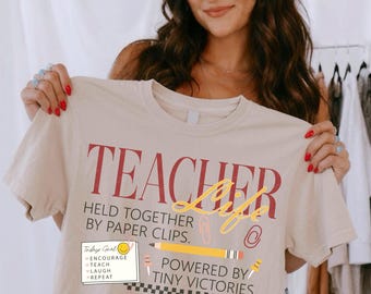 May include: A beige short-sleeve t-shirt featuring the red text TEACHER LIFE, the phrase HELD TOGETHER BY PAPER CLIPS, and POWERED BY TINY VICTORIES. The design includes a yellow pencil, paper clips, and a note with a smiley face and a checklist.