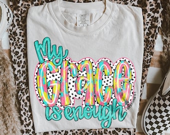 May include: A cream-colored Comfort Colors t-shirt with the text "My Grace is enough" in a colorful, layered design. The shirt is paired with leopard print pants and a pair of checkered slip-on shoes.