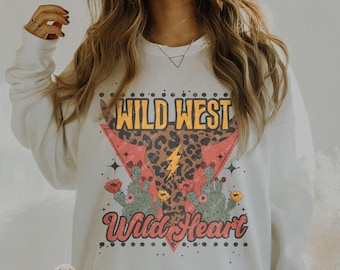 Peut inclure: Sweat-shirt blanc avec un imprimé graphique d'une tête de taureau à imprimé léopard avec un éclair et des cactus. Le texte "Wild West Wild Heart" est imprimé sous la tête de taureau.