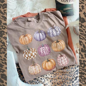 May include: A tan t-shirt featuring a watercolor design of nine pumpkins in various colors and patterns. The pumpkins include orange, purple, pink, and white, with designs such as polka dots, stars, and checkered patterns. The shirt is part of a fall-themed collection.