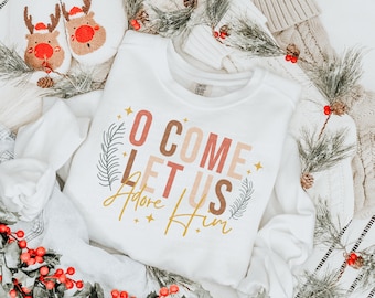 Puede incluir: Sudadera blanca con el texto "O Come Let Us Adore Him" en letras rosa, marrón y doradas con un diseño de hoja dorada.