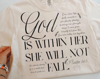 May include: A cream-colored short-sleeve t-shirt featuring black text that reads: God IS WITHIN HER SHE WILL NOT FALL. Additional text includes: Even when life feels uncertain, she stands steady, knowing her strength doesn't come from herself but from the One who holds her together. She stands steady in a world that tries to shake her. Psalm 46:5, along with small leaf and cross graphics.