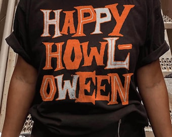 May include: A black short-sleeved t-shirt featuring the text HAPPY HOWLOWEEN printed in a distressed, alternating orange and white font. The graphic is centred on the chest, utilising a bold, blocky typeface with a weathered, vintage texture.
