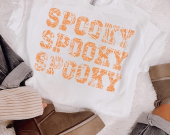 Może przedstawiać: Biała koszulka z napisem "SPOOKY" w kolorze pomarańczowym, powtórzonym trzykrotnie w stylu vintage. Na zdjęciu brązowe buty i dżinsy, sugerujące jesienny lub halloweenowy motyw.