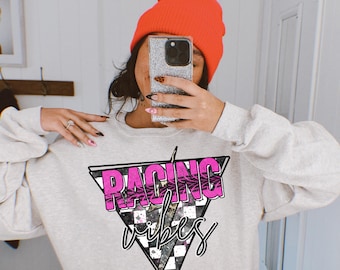 Op de afbeelding: Een grijs sweatshirt met een roze driehoekige afbeelding waarop in een retro-lettertype "Racing Vibes" staat. De driehoekige afbeelding heeft een geruit patroon en een bliksem.