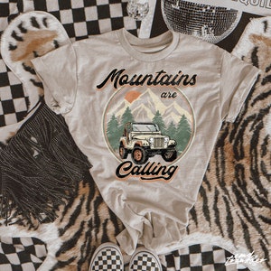 May include: Beige t-shirt with a circular graphic featuring a jeep in front of mountains and trees, with the text "Mountains are Calling". The design includes an orange sun and a green border.