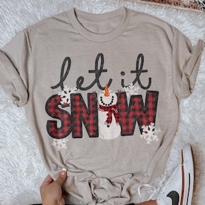 May include: Beige long-sleeve t-shirt with the phrase "let it snow" in black script and red plaid block letters. A snowman with a carrot nose and red scarf is in the "O" of "SNOW". White snowflakes are scattered around the text.