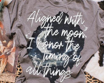 May include: A charcoal gray short-sleeve t-shirt featuring a central graphic of a moonlit night sky with stars and clouds. The white cursive text reads: Aligned with the moon, I honor the timing of all things. The shirt has a crew neckline.