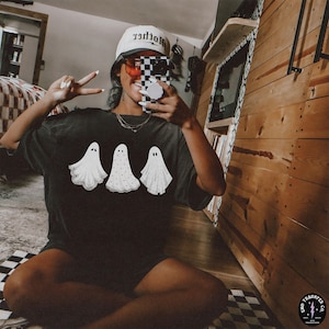 May include: A black t-shirt with a graphic of three white ghosts. The ghosts are wearing a simple, dotted pattern. The person wearing the shirt is holding a phone with a black and white checkered case. The shirt has the text "Mother" on the hat.