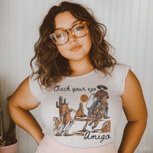 May include: A white t-shirt with a graphic of a cowboy riding a horse. The graphic includes the text "Check your ego" and "Amigo".