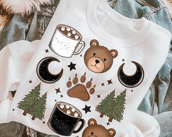 May include: A white crewneck sweatshirt featuring a graphic print of two brown bear faces, two mugs of hot cocoa with marshmallows, two crescent moons with starry patterns, two clusters of green pine trees, a bear paw print, and small stars.