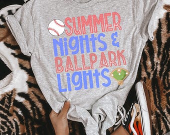May include: A heather gray t-shirt with the text "SUMMER NIGHTS & BALLPARK LIGHTS" in red and blue. The shirt features a baseball and a baseball field graphic. The shirt is paired with black and white sneakers with an orange flame design.