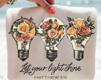 May include: A light beige tote bag featuring three light bulb designs filled with roses in shades of pink, yellow, and red. The text "Let your light shine" and "Matthew 5:16" are printed in black.