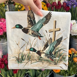 May include: A white square decorative pillow featuring two flying ducks with green and brown feathers, a wooden cross, and reeds. The ducks are depicted above a body of water. The pillow is held by a person.