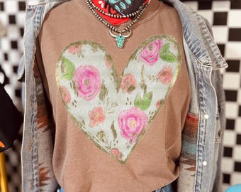 Puede incluir: Una camiseta marrón de manga larga con un diseño de corazón lleno de rosas rosas y hojas verdes. El corazón está delineado en dorado. La camiseta se combina con una chaqueta vaquera, una bandana y un collar de toro turquesa.