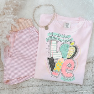 May include: A light pink t-shirt with the word "LOVE" in a colorful, whimsical design, featuring a pencil, apple, and notebook elements. The shirt also has the text "Let all that you do be done in" and "1 Cor 16:14". A silver necklace and pink gingham shorts are also in the image.