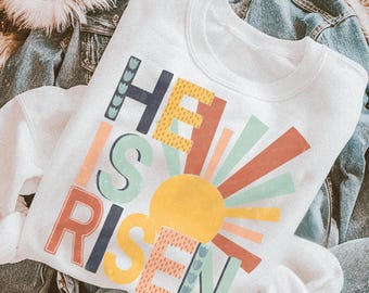 May include: A white crewneck sweatshirt featuring the text HE IS RISEN in a multi-colored, distressed font. The design includes a yellow sun graphic with radiating beams in shades of orange, green, and yellow, accented with small blue floral motifs.