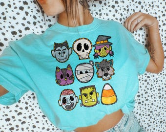 May include: A light blue t-shirt featuring a collection of Halloween-themed cartoon faces. The design includes a vampire, skull, witch, cat, mummy, wolf, zombie, Frankenstein's monster, and candy corn. The shirt is made of a soft material.