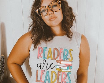 May include: A woman wearing a white tank top with the text "Readers are Leaders" in colorful letters and a stack of books underneath the text. She is wearing tortoise shell glasses and has long brown hair.
