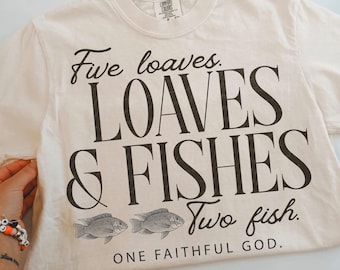 May include: A cream-colored short-sleeve t-shirt featuring black text that reads Five loaves. LOAVES & FISHES Two fish. ONE FAITHFUL GOD. The design includes two small fish illustrations positioned between the lines of text on the front.