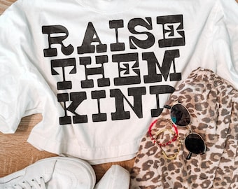 May include: A white t-shirt featuring the black text RAISE THEM KIND in a bold, blocky font. The shirt is paired with leopard print shorts, white trainers, a pair of round sunglasses, and two beaded bracelets with red and yellow accents.