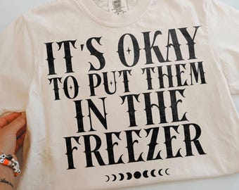 May include: A cream-colored short-sleeve t-shirt featuring black text that reads IT'S OKAY TO PUT THEM IN THE FREEZER. Below the text is a horizontal row of black moon phase icons. The shirt has a crew neckline and a Comfort Colors brand label.