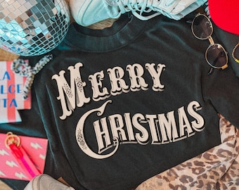 May include: A black long-sleeve shirt featuring the white text Merry Christmas in a stylized font. The shirt is surrounded by a silver disco ball, light blue high-top sneakers, round sunglasses, a leopard print fabric, and a pink keychain.