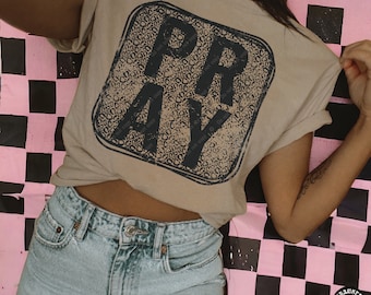 May include: A tan t-shirt with a black square graphic that says "PRAY" in a leopard print pattern. The shirt is being worn by a person with a tattoo on their arm.
