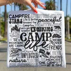 May include: White t-shirt with a black graphic design featuring camping-themed words and illustrations, including "Camp Life," "Adventure Awaits," and "Mountains are Calling." The design includes a campfire, stars, and a bear.