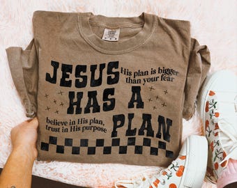 May include: A brown t-shirt with the text "JESUS HAS A PLAN" in black. The text "His plan is bigger than your fear" is also in black. The text "believe in His plan, trust in His purpose" is in black and smaller font.