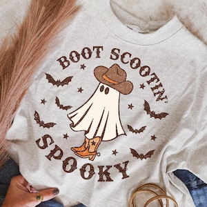 May include: A light gray crewneck t-shirt featuring a graphic of a ghost wearing a brown cowboy hat and orange cowboy boots. The text "BOOT SCOOTIN' SPOOKY" is printed in brown around the ghost, surrounded by small brown bats and stars.