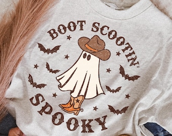 Może przedstawiać: Jasnoszary t-shirt z okrągłym dekoltem z grafiką ducha w brązowym kowbojskim kapeluszu i pomarańczowych kowbojkach. Napis "BOOT SCOOTIN' SPOOKY" jest nadrukowany na brązowo wokół ducha, w otoczeniu małych brązowych nietoperzy i gwiazdek.