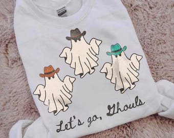 May include: A white crewneck sweatshirt featuring a graphic of three ghosts wearing cowboy hats in brown, black, and teal. The text "Let's go, Ghouls" is printed in a black cursive font below the ghosts. The garment has a standard long-sleeve design.