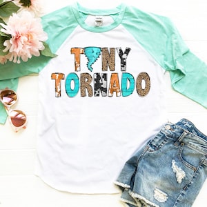 May include: A white and turquoise raglan t-shirt with the text "Tiny Tornado" in a colorful, distressed font. The text is decorated with cowhide, leopard, and turquoise patterns.
