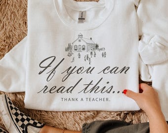 May include: A white crewneck sweatshirt featuring a black line drawing of a schoolhouse with children playing in front. The text reads, "If you can read this... THANK A TEACHER." in a combination of cursive and block lettering on the front.