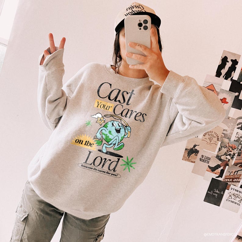 Puede incluir: Una sudadera gris claro con el texto "Cast Your Cares on the Lord" y un gr&aacute;fico de la tierra de dibujos animados. La sudadera tiene mangas largas y cuello redondo. La persona de la imagen lleva una gorra de b&eacute;isbol y sostiene un tel&eacute;fono.