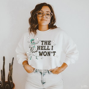Puede incluir: Sudadera color crema con un gr&aacute;fico de vaquera y el texto "THE HELL I WON'T" en negro. La vaquera lleva un sombrero y botas turquesas, sosteniendo un lazo. Estrellas rodean el texto y el gr&aacute;fico.